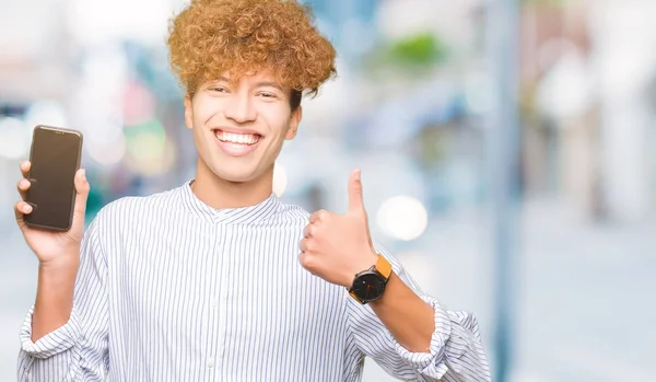 年轻英俊的男人显示智能手机屏幕快乐与灿烂的笑容做确定的迹象 用手指竖起大拇指 优秀的标志 — 图库照片
