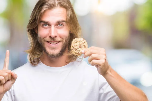 若いハンサムな男長い髪を食べるチョコレート クッキーを分離アイデアや質問 ナンバーワンの幸せそうな顔で指を指していると驚かれる背景 — ストック写真