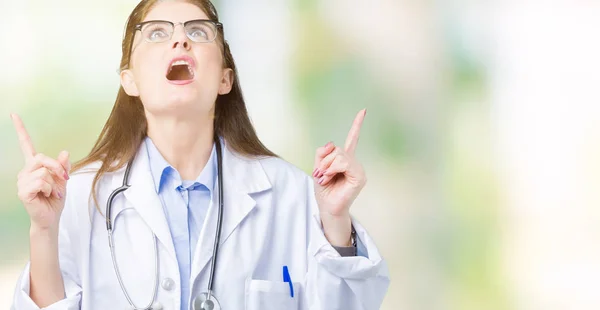 中年成熟的医生妇女穿着医疗外套在孤立的背景惊讶和惊讶地抬起头来 用手指和举起的手臂指指点点 — 图库照片