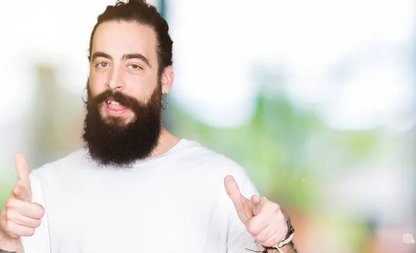 Young hipster man with long hair and beard wearing casual white t-shirt pointing fingers to camera with happy and funny face. Good energy and vibes.