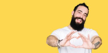 Uzun saç ve sakal casual beyaz t-shirt aşık kalp simgesi ve elleriyle şekil gösteren gülümseyen giyen genç hipster adam. Romantik kavramı.