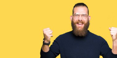 Gözlük ve balıkçı yaka kazak kutluyor giyen genç sarışın adam şaşırttı ve başarı için kaldırdı kollarını açarak hayret ve gözleri açık. Kazanan kavramı.