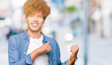 Afro saçlı kot giyen genç yakışıklı adam kendinden emin gülümseyen işaretleme için arkasından el ve başparmak yukarıya, ceket