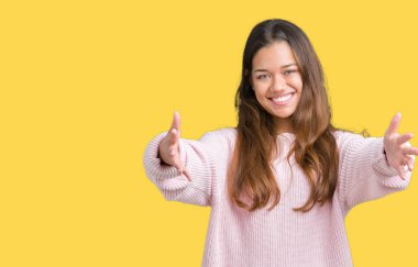 Kamera ile gülümseyen bakarak izole arka plan üzerinde pembe kış kazak giyen genç güzel esmer kadın kolları sarılmak için açın. Neşeli ifade kucaklayan mutluluk.