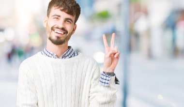 Mutlu yüz zafer işareti yaparken kameraya göz kırpıyor ile gülümseyen izole arka plan üzerinde kış kazak giyen genç yakışıklı adam. İki numaralı.