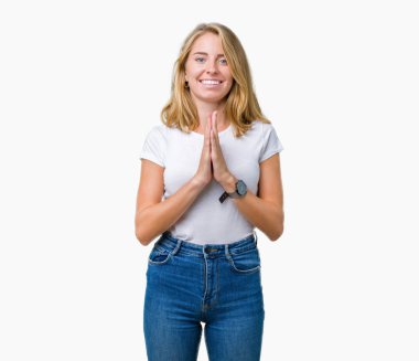 Ellerle birlikte AF emin gülümseyen için soran dua izole arka plan üzerinde rahat beyaz t-shirt giyen güzel genç kadın.