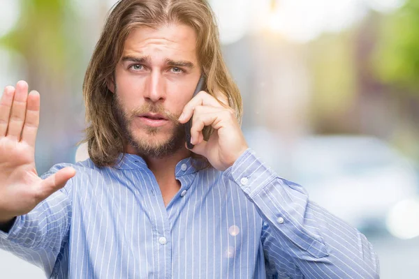 年轻的英俊男子与长头发在孤立的背景说话在电话中张开手做停止标志与严肃和自信的表情 防御手势 — 图库照片