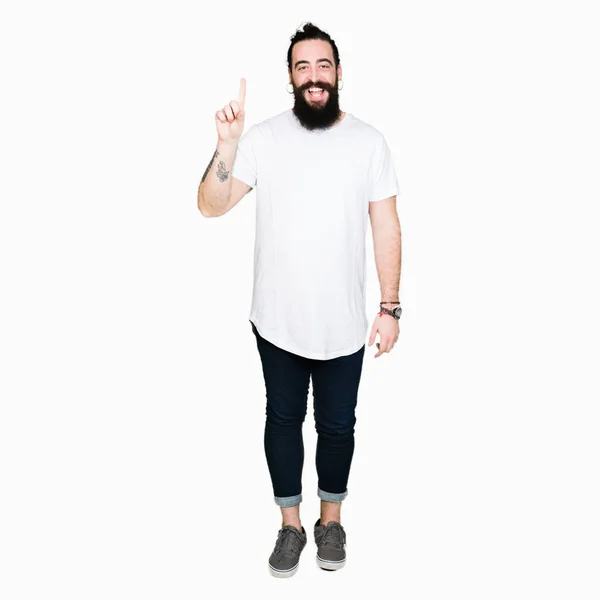年轻的嬉皮士男子长头发和胡子穿着休闲白色 T恤显示和手指第一的指向 同时微笑着自信和快乐 — 图库照片