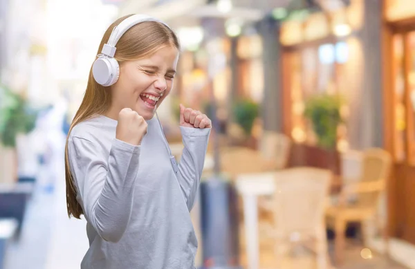 年轻美丽的女孩戴着耳机听音乐在孤立的背景非常高兴和兴奋做优胜者手势举起双臂 微笑着 尖叫着要成功 庆祝理念 — 图库照片