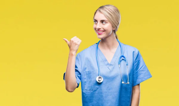 年轻美丽的金发医生外科医生护士妇女在孤立的背景微笑与愉快的面孔看 用拇指指向边 — 图库照片