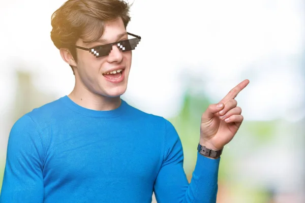 Young man wearing funny thug life glasses over isolated background with a big smile on face, pointing with hand and finger to the side looking at the camera.