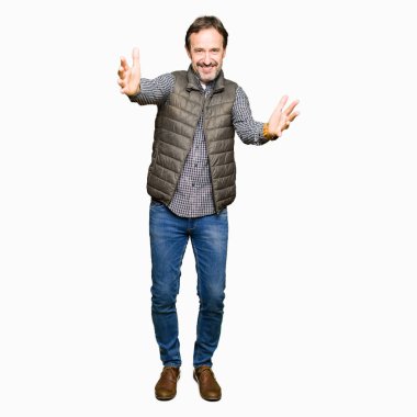 Orta yaş yakışıklı adam sarılmak için kollarını açarak gülümseyerek kameraya bakarak kış yelek giyiyor. Neşeli ifade kucaklayan mutluluk.
