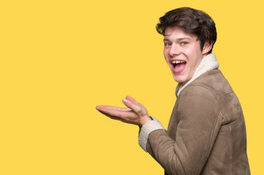 Genç yakışıklı adam giyen kış ceket izole arka plan üzerinde el ve reklam mutlu ve kendinden emin gülümseyen sunulması avuç, yanında işaret