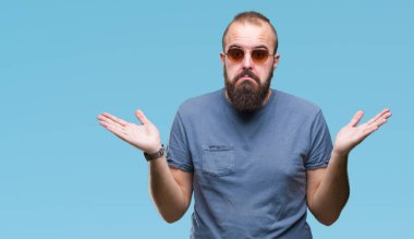 Genç beyaz hipster adam izole arka plan clueless ve şaşkın bir ifadeyle kollar ve eller kaldırdı üzerinde güneş gözlüğü takıyor. Şüphe kavramı.