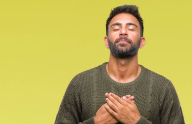 Yetişkin İspanyol adam ile gülümseyen izole arka plan üzerinde kış kazak giyen kapalı gözler ve yüzünde minnettar jest ile göğüs üzerinde eller. Sağlık kavramı.