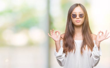 Genç Asyalı kadın giyiyor güneş gözlüğü izole arka plan üzerinde dinlenmek ve gülümseyen gözlerle kapalı parmak jestle meditasyon yapıyor. Yoga kavramı.