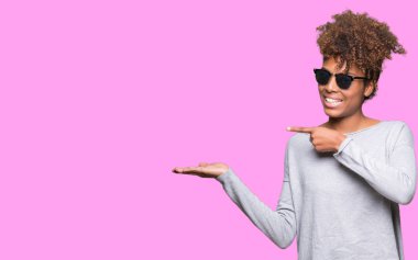 Güzel genç Afro-Amerikan kadın hayrete ve el ile sunulması ve parmak ile işaret ederken kameraya gülümseyen izole arka plan üzerinde güneş gözlüğü takıyor.