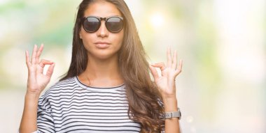 Genç güzel Arap kadın giyiyor güneş gözlüğü izole arka plan üzerinde dinlenmek ve gülümseyen gözlerle kapalı parmak jestle meditasyon yapıyor. Yoga kavramı.