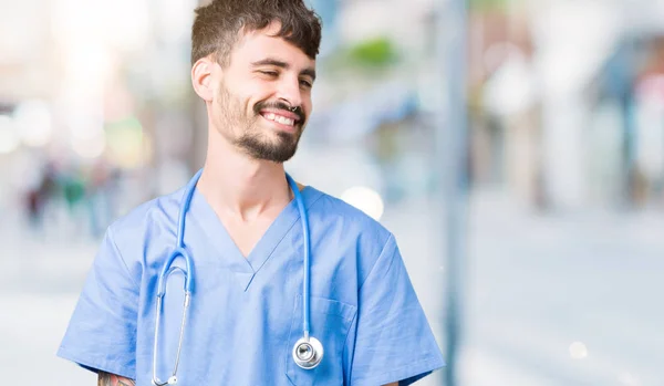 Young handsome nurse man wearing surgeon uniform over isolated background looking away to side with smile on face, natural expression. Laughing confident.