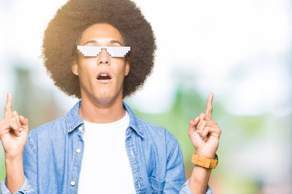 Young african american man with afro hair wearing thug life glasses amazed and surprised looking up and pointing with fingers and raised arms.