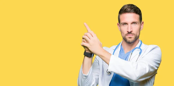 Handsome doctor man wearing medical uniform over isolated background Holding symbolic gun with hand gesture, playing killing shooting weapons, angry face
