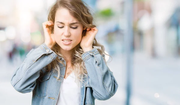 美しい若い金髪女性の騒々しい音楽のノイズの腹式指で耳を覆う孤立した背景にデニム ジャケットを着ています 聴覚障害者の概念 — ストック写真
