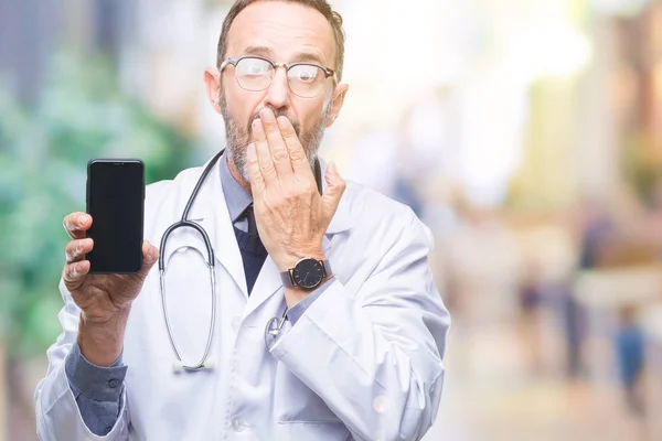 Middle age senior hoary doctor man showing smartphone screen over isolated background cover mouth with hand shocked with shame for mistake, expression of fear, scared in silence, secret concept