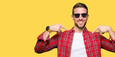 Yakışıklı adam yüzünde gülümseme ile kendinden emin görünüyorsun, kendini gururlu ve mutlu parmakları ile işaret moda güneş gözlüğü takıyor.