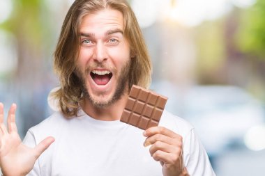 Uzun saç beslenme ile yakışıklı delikanlı büyük gülümseme ile çığlık zaferini kutluyor ve ellerini kaldırdı izole arka plan çok mutlu ve heyecanlı, kazanan ifade üzerinde çikolata bar