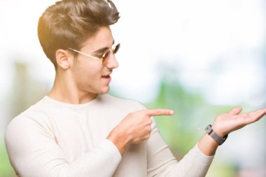 Yakışıklı delikanlı hayrete ve el ile sunulması ve parmak ile işaret ederken kameraya gülümseyen izole arka plan üzerinde güneş gözlüğü takıyor.