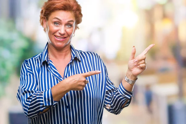 Atrractive senior caucasian redhead woman over isolated background smiling and looking at the camera pointing with two hands and fingers to the side.