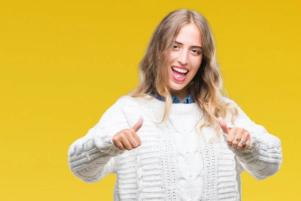 Beautiful young blonde woman wearing winter sweater over isolated background approving doing positive gesture with hand, thumbs up smiling and happy for success. Looking at the camera, winner gesture.