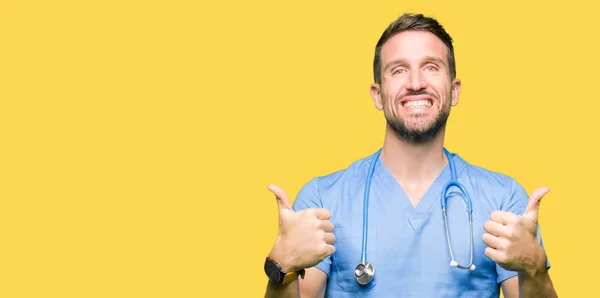 帅气的医生在孤立的背景成功标志上穿上医疗制服做积极的手势 竖起大拇指微笑和快乐 带着欢快的表情 获胜者的手势看着镜头 — 图库照片