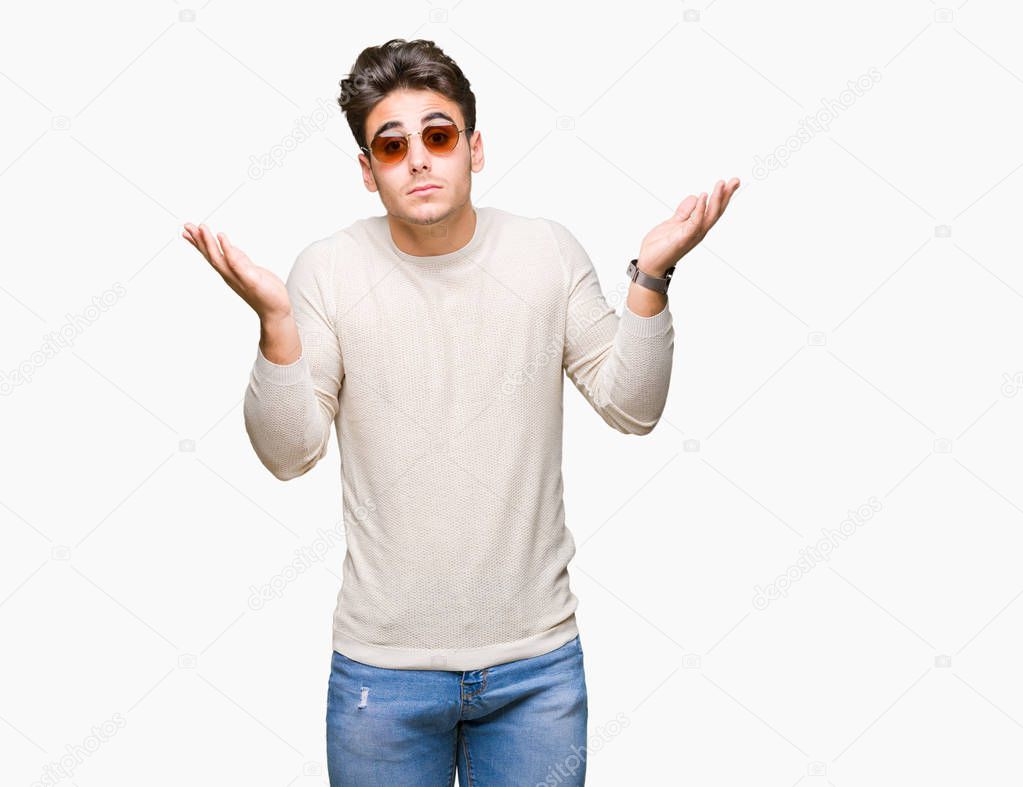 Young handsome man wearing sunglasses over isolated background clueless and confused expression with arms and hands raised. Doubt concept.
