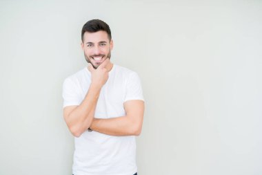 Emin çapraz kollar ve çeneme kaldırdı el ile gülümseme ile kameraya bakarak izole arka plan üzerinde rahat beyaz t-shirt giyen genç yakışıklı adam. Pozitif düşünce.