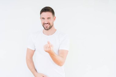 Beckoning gel burada mutlu ve gülümseyerek el davet ile jest izole arka plan üzerinde rahat beyaz t-shirt giyen genç yakışıklı adam