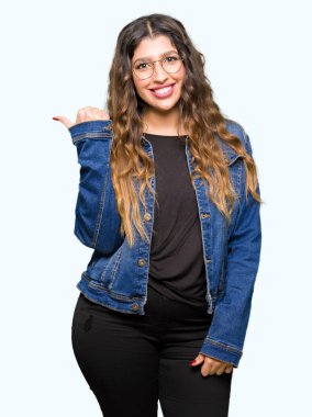 Gözlük ve seyir ve başparmak yanında yukarıyı mutlu yüz ile gülümseyen denim ceket giyen genç güzel kadın.