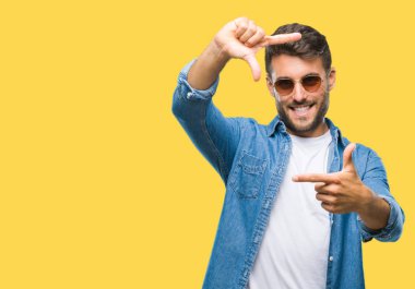Genç yakışıklı adam elleriyle yapım çerçeve ve parmakları ile mutlu yüz gülümseyen izole arka plan üzerinde güneş gözlüğü takıyor. Yaratıcılık ve Fotoğrafçılık kavramı.