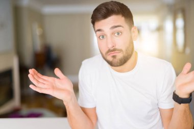 Genç yakışıklı giyiyor rahat beyaz t-shirt ev clueless ve şaşkın bir ifadeyle kollar ve eller kaldırdı, adam. Şüphe kavramı.