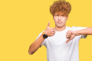 Yakışıklı genç afro saçlı casual beyaz tişört yapıyor başparmak yukarı ve aşağı giyiyor, anlaşmazlık ve anlaşma ifade. Deli çatışma