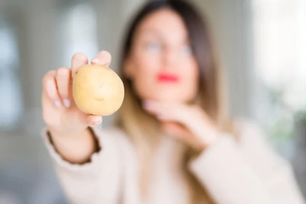 年轻美丽的女人拿着新鲜的土豆在家里严肃的脸思考问题 很困惑的想法 — 图库照片