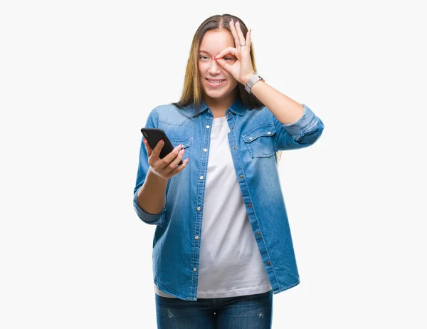 年轻美丽的高加索妇女发短信发送消息使用智能手机在孤立的背景与快乐的脸微笑做确定的标志与眼睛看通过手指 — 图库照片