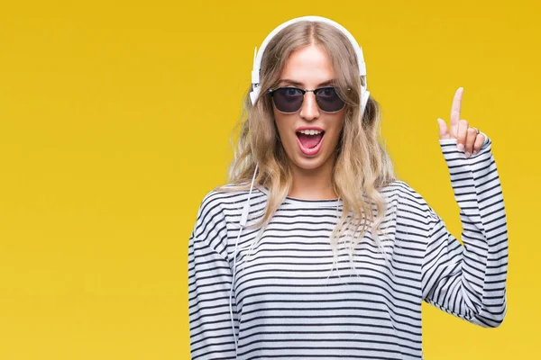Başarılı fikirle parmağı yukarıyı izole arka plan üzerinde kulaklık ve güneş gözlüğü giyen güzel genç sarışın kadın. Çıktıktan ve mutlu. Bir numara.