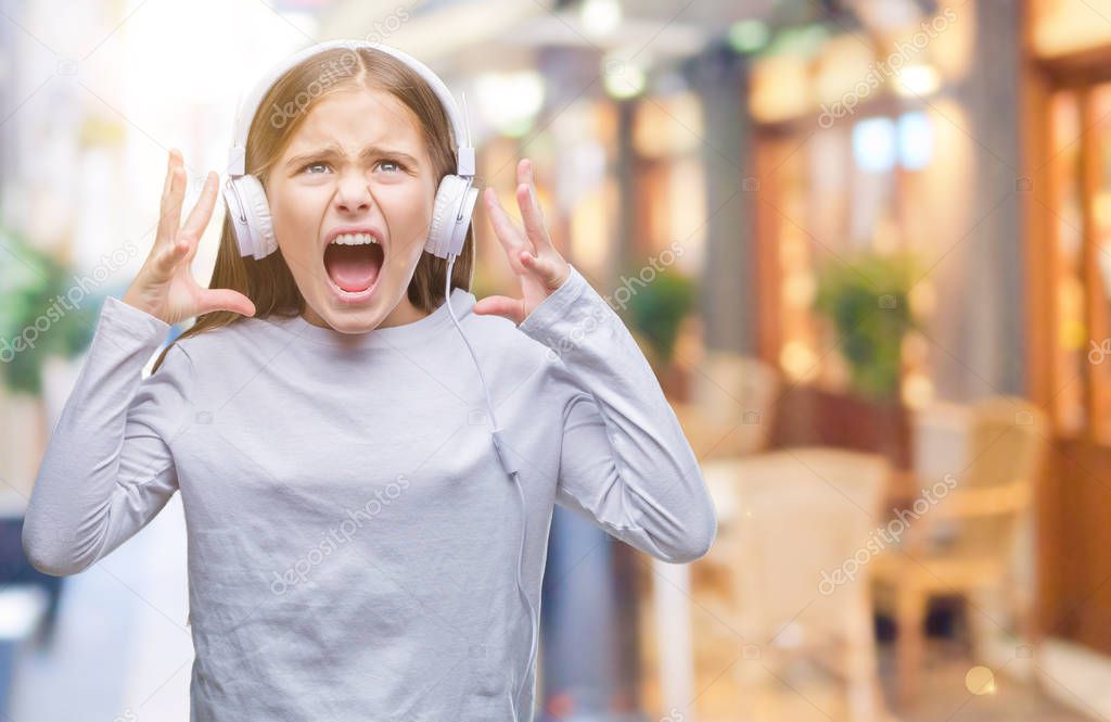 Young beautiful girl wearing headphones listening to music over isolated background crazy and mad shouting and yelling with aggressive expression and arms raised. Frustration concept.