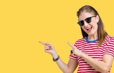 Orta yaş Olgun kadın gülümseyerek ve iki elleri ve parmakları ile tarafına işaret kamera bakarak izole arka plan üzerinde güneş gözlüğü takıyor.