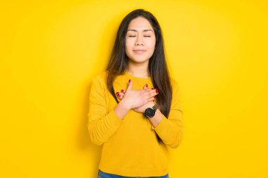 Sarı izole arka plan ile gülümseyen güzel esmer kadın kapalı gözler ve yüzünde minnettar jest ile göğüs üzerinde eller. Sağlık kavramı.