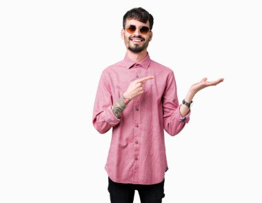 Yakışıklı delikanlı hayrete ve el ile sunulması ve parmak ile işaret ederken kameraya gülümseyen izole arka plan üzerinde güneş gözlüğü takıyor.