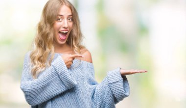 Hayret ve el ile sunulması ve parmak ile işaret ederken kameraya gülümseyen izole arka plan üzerinde kış kazak giyen güzel genç sarışın kadın.