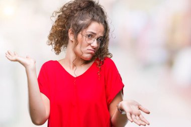 Güzel esmer kıvırcık saçlı Genç kız üzerinde gözlük takan arka plan clueless ve şaşkın bir ifadeyle kollar ve eller kaldırdı izole. Şüphe kavramı.