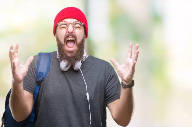 Üzerinde kırmızı yün kap ve sırt çantası giyen genç hipster adam arka plan deli izole ve deli bağırarak ve agresif ifade ve silah kaldırdı bağırıyor. Hayal kırıklığı kavramı.